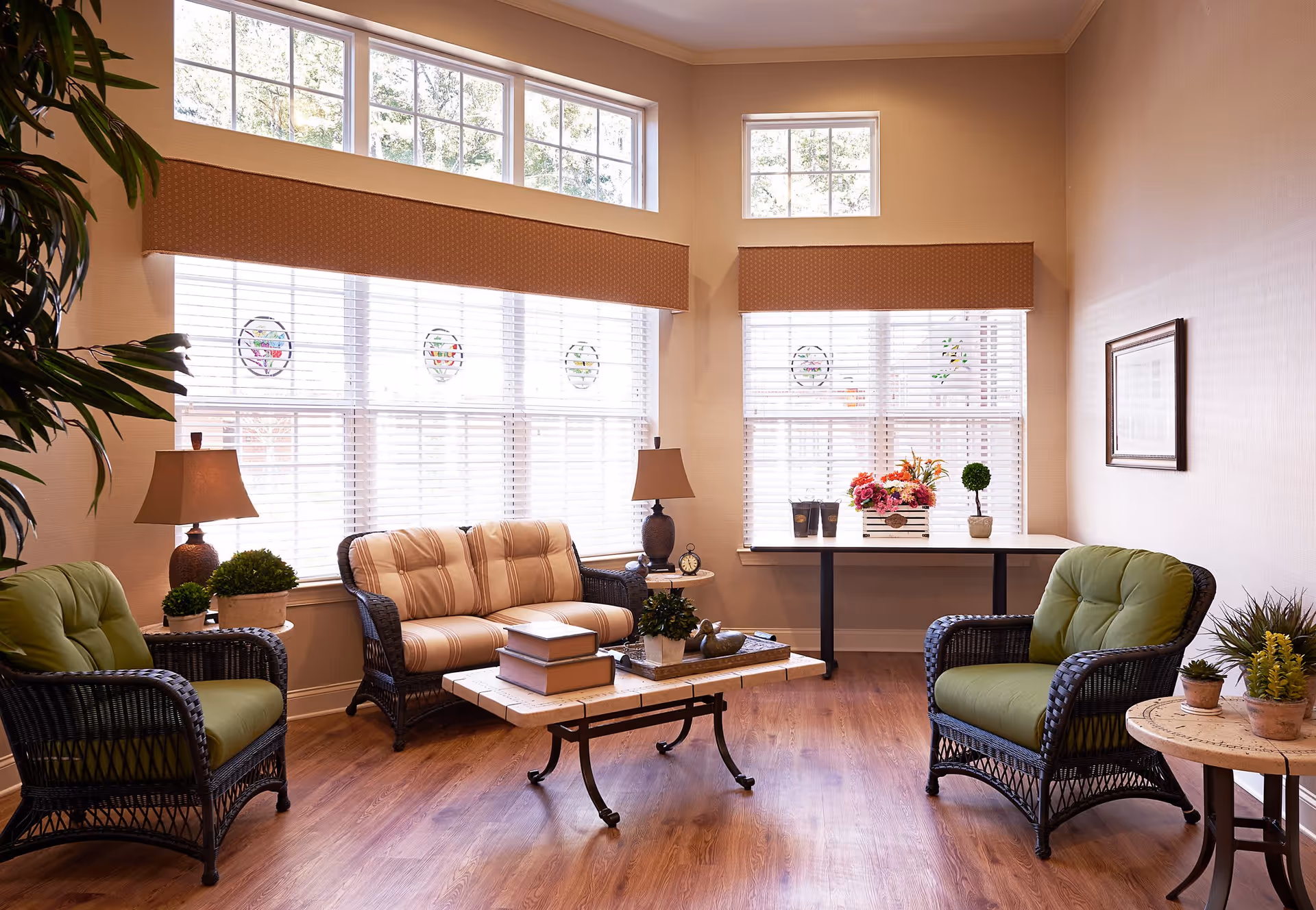 A cozy living room with large windows featuring white blinds and decorative stained glass accents. The room has a wooden floor, two green cushioned wicker armchairs, a striped cushioned wicker loveseat, a coffee table with books and plants, side tables with lamps and plants, and a table with a flower arrangement and decorative items near the window.