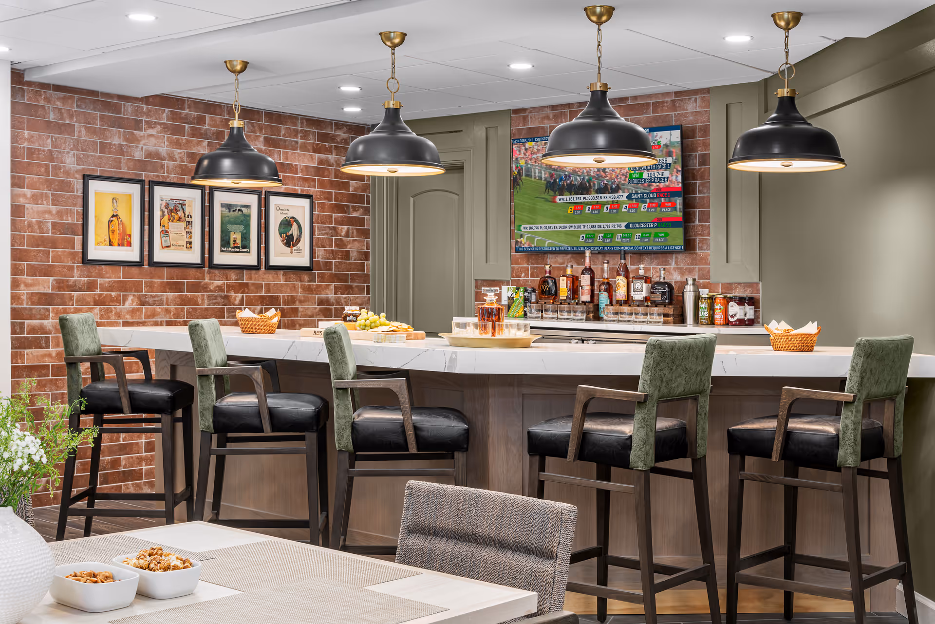 A modern bar area with a white marble countertop and six high chairs with green backs and black seats. The back wall is covered with red brick tiles and decorated with four framed pictures and a mounted flat-screen TV displaying a sports event. Three black pendant lights hang from the ceiling above the bar. Various bottles and glasses are arranged on the counter and shelves behind the bar. In the foreground, part of a dining table with bowls of snacks and a white vase with flowers is visible.