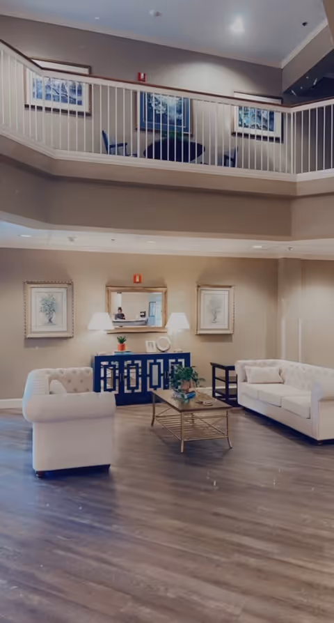 A spacious and well-lit living room area with two white tufted sofas facing each other, a glass coffee table with a plant on it, a dark blue sideboard with two lamps and decorative items, framed artwork on the walls, and a balcony railing above overlooking the space.