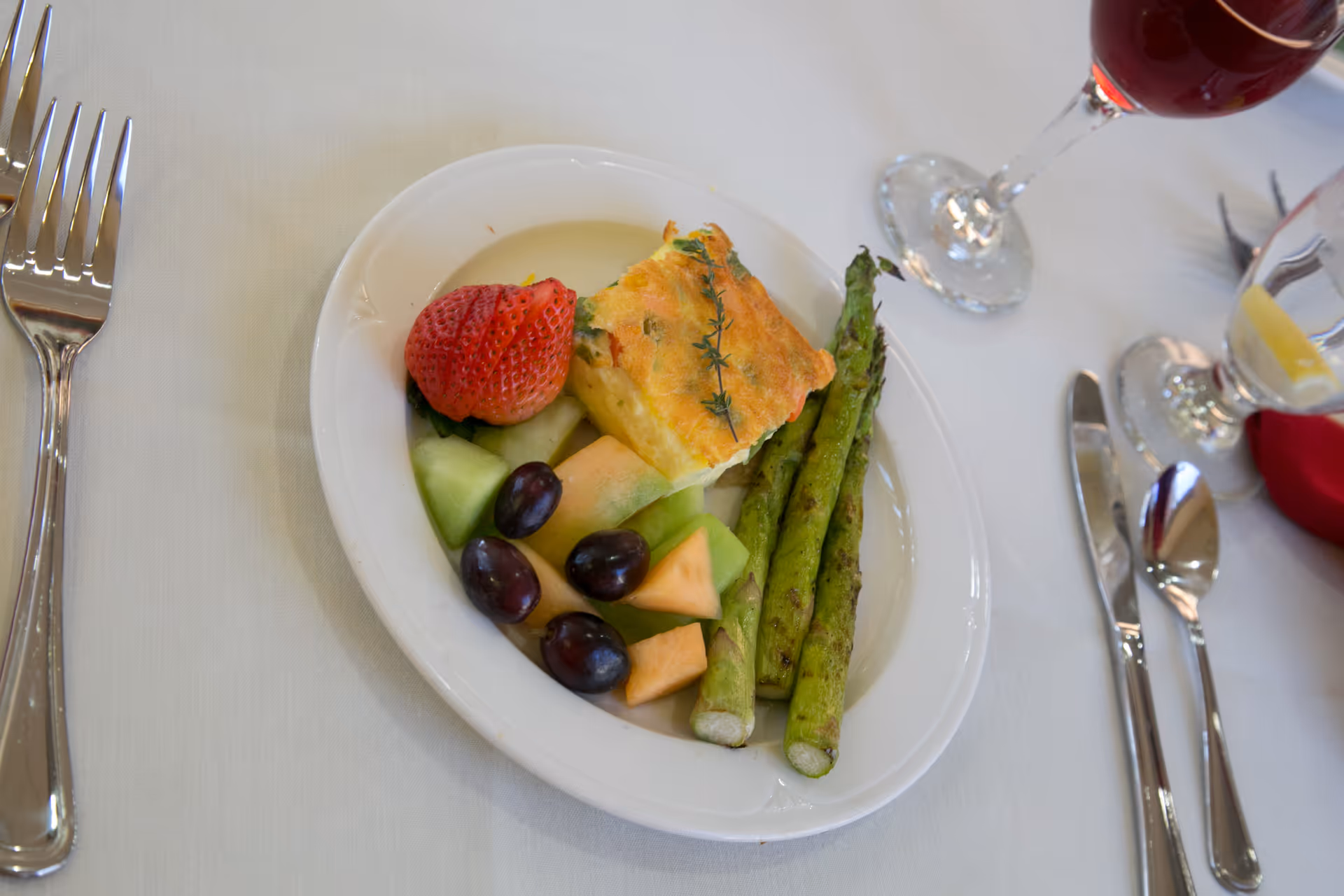 A white plate with a serving of grilled asparagus, a piece of baked egg dish garnished with a sprig of thyme, and a small assortment of fresh fruit including a strawberry, cantaloupe, honeydew melon, and grapes. The plate is set on a white tablecloth with silverware and glasses of water and red wine nearby.