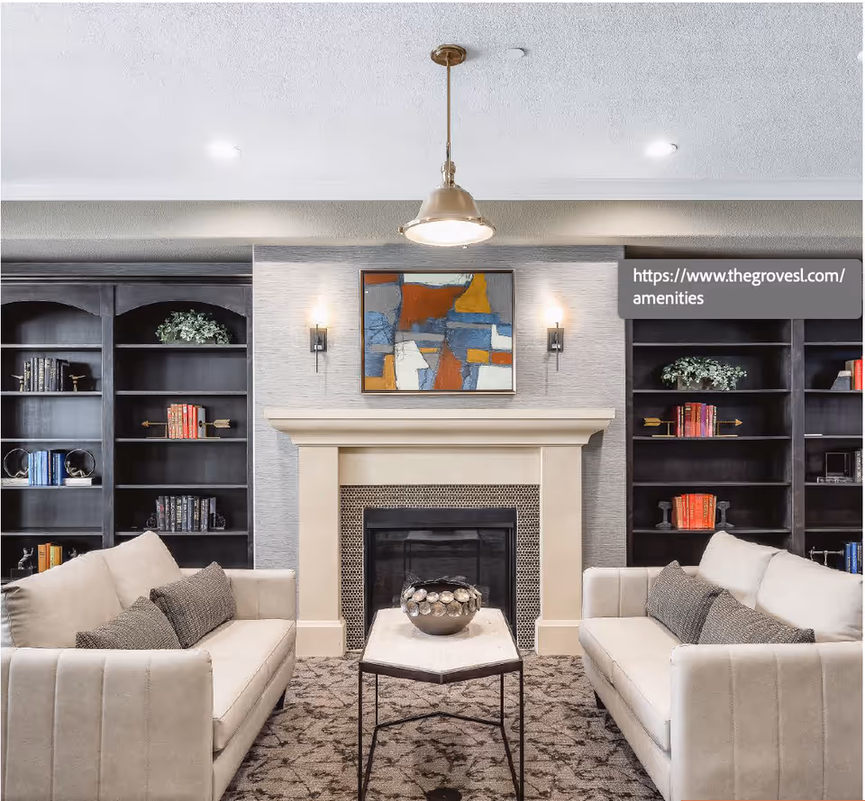 A cozy living room with two beige sofas facing each other across a small coffee table. In the center is a fireplace with a decorative bowl on the table. Behind the fireplace are built-in dark wood bookshelves with books and decorative items. Above the fireplace hangs an abstract painting with blue, orange, white, and yellow colors. The room is softly lit by wall sconces and a hanging ceiling light.