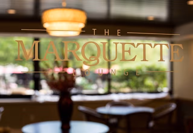View through a glass window or door with the words 'The Marquette Lounge' etched on it, showing a blurred interior with tables, chairs, a vase with flowers, and a hanging light fixture, with greenery visible outside the window.