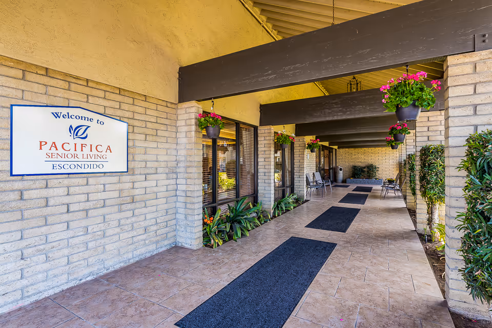 Covered entrance walkway with hanging flower pots, seating, and a 'Pacifica Senior Living Escondido' sign on a brick wall.