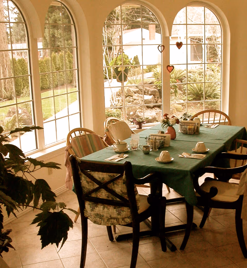 A dining area with a rectangular table covered with a green tablecloth, set with cups, saucers, utensils, and small flower arrangements. The room has large arched windows showing a garden with greenery and rocks outside. Several chairs with cushions surround the table.