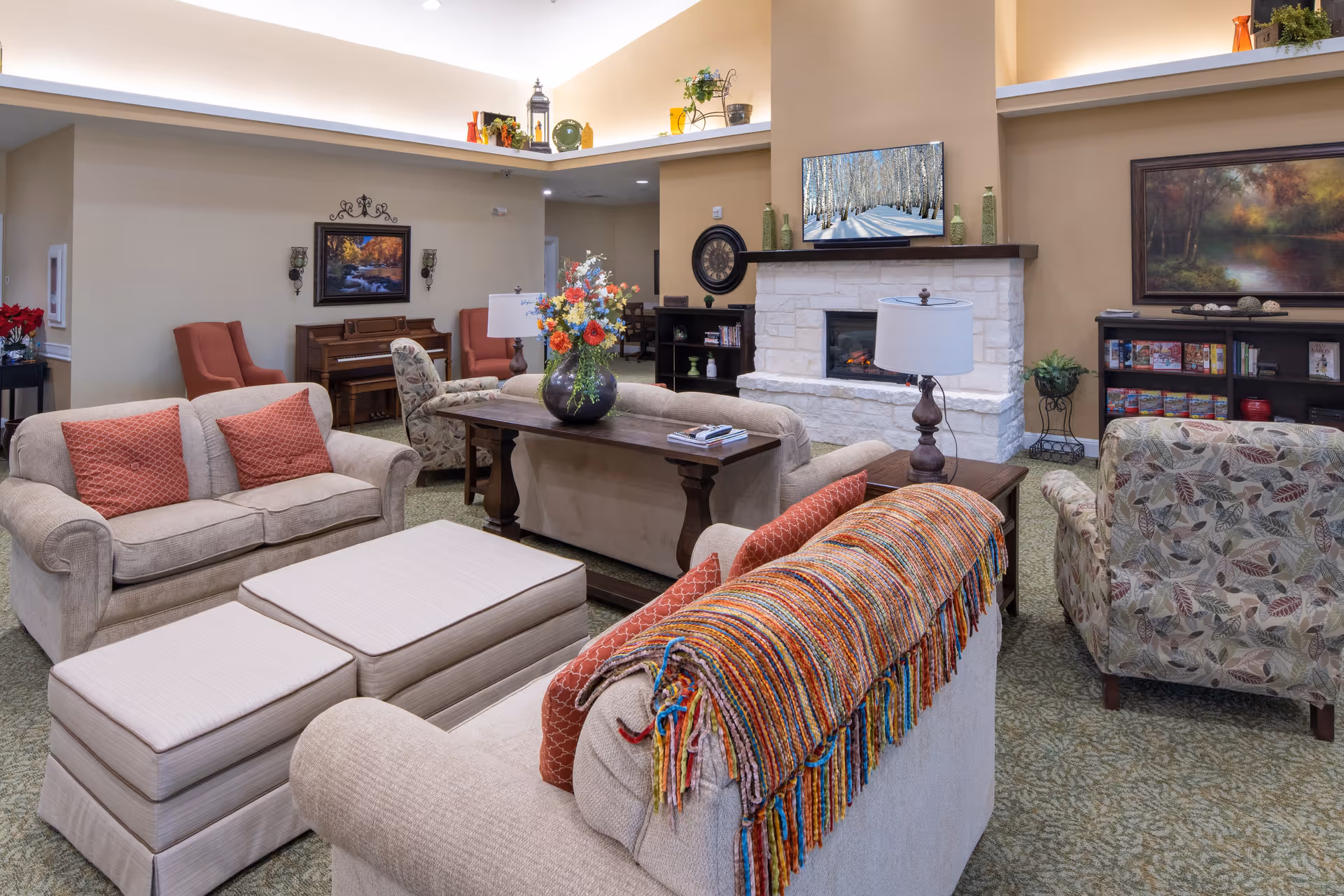 A cozy living room in an assisted living facility featuring beige sofas with red cushions, a colorful throw blanket, a floral armchair, a wooden coffee table with a vase of flowers, a white stone fireplace with a TV mounted above it, a piano in the background, and bookshelves with books and decorative items.