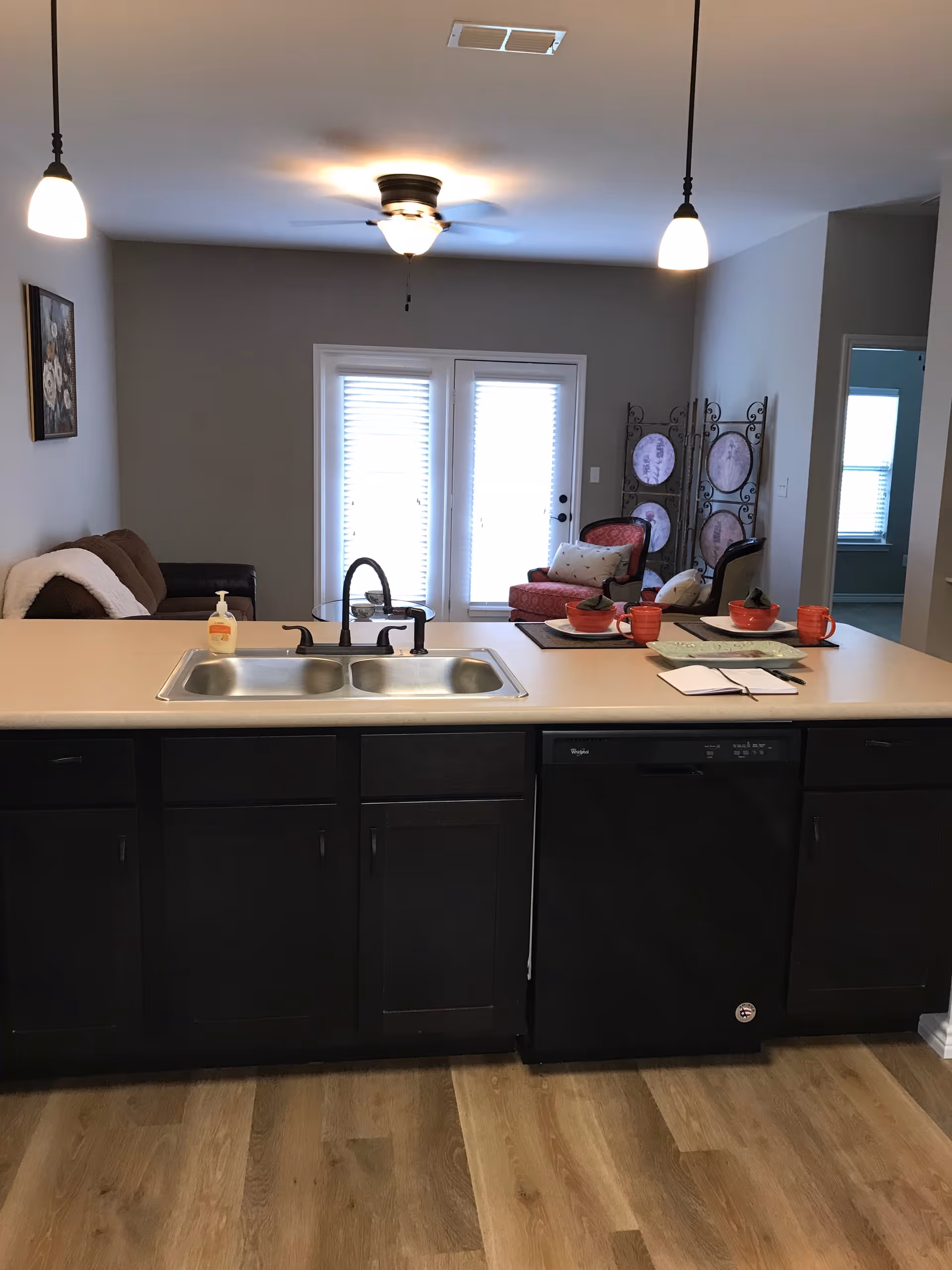 View of a kitchen island with a double sink and black cabinets, a dishwasher, and a countertop with red bowls and mugs. In the background, there is a living area with a brown couch, a red armchair, a decorative folding screen, and French doors with blinds. The ceiling has a fan with a light and two hanging pendant lights.