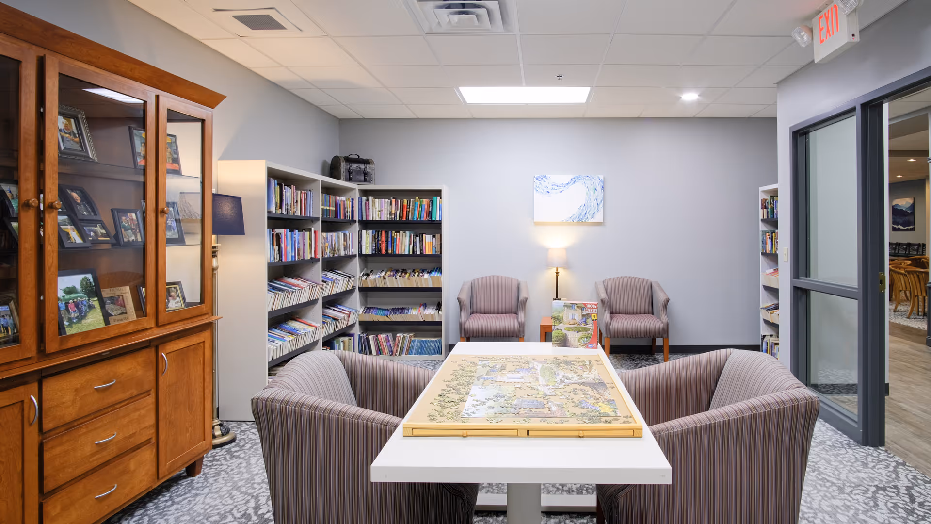 A cozy reading and activity room in a senior living facility featuring a white table with a puzzle on it, surrounded by four striped armchairs. In the background, there are bookshelves filled with books, a wooden cabinet with framed photos, a floor lamp, and two additional armchairs with a small table and lamp between them. The room has a patterned carpet and neutral-colored walls with a piece of artwork hanging above the chairs.