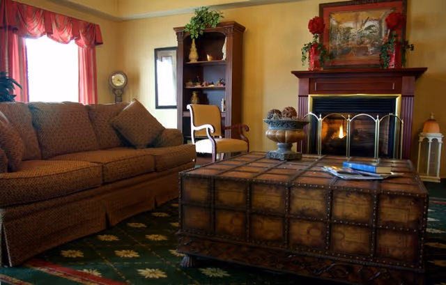 A cozy living room with a patterned brown sofa, a wooden armchair with yellow upholstery, and a large wooden coffee table with a decorative bowl and magazines on it. There is a fireplace with a fire burning, flanked by red floral decorations and a framed painting above it. A wooden bookshelf with decorative items is in the background, and a window with red curtains lets in natural light.