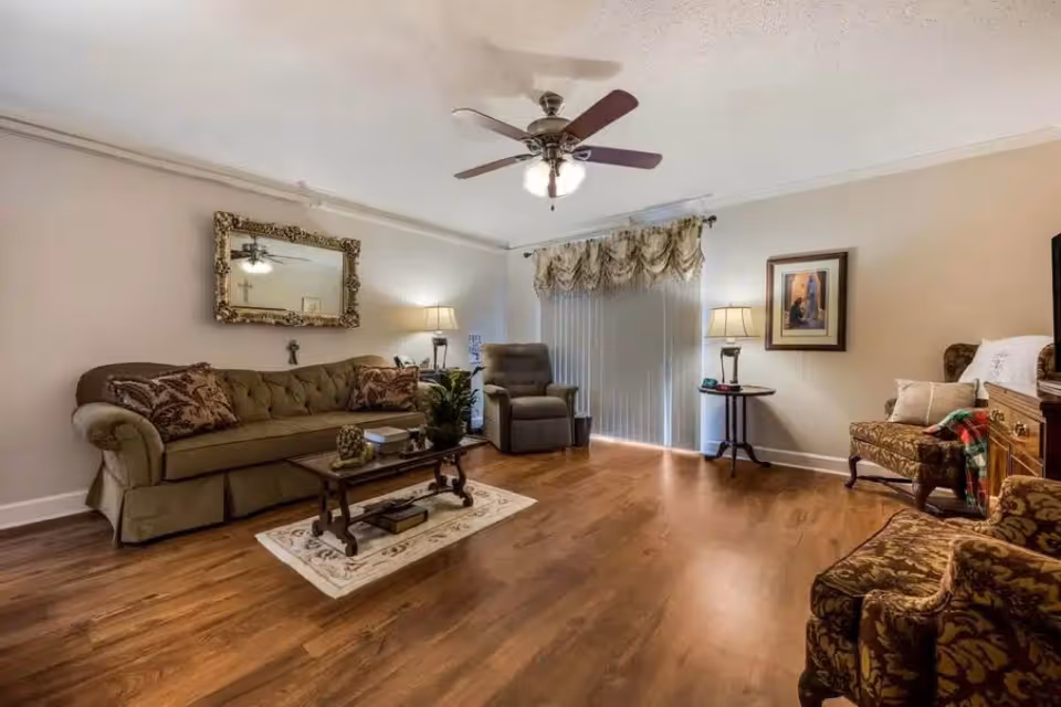 A cozy living room with wooden flooring, a ceiling fan with lights, a large olive green sofa with patterned cushions, a glass coffee table on a small rug, a recliner chair, two patterned armchairs, two side tables with lamps, a large ornate mirror on the wall, and vertical blinds covering a sliding glass door.