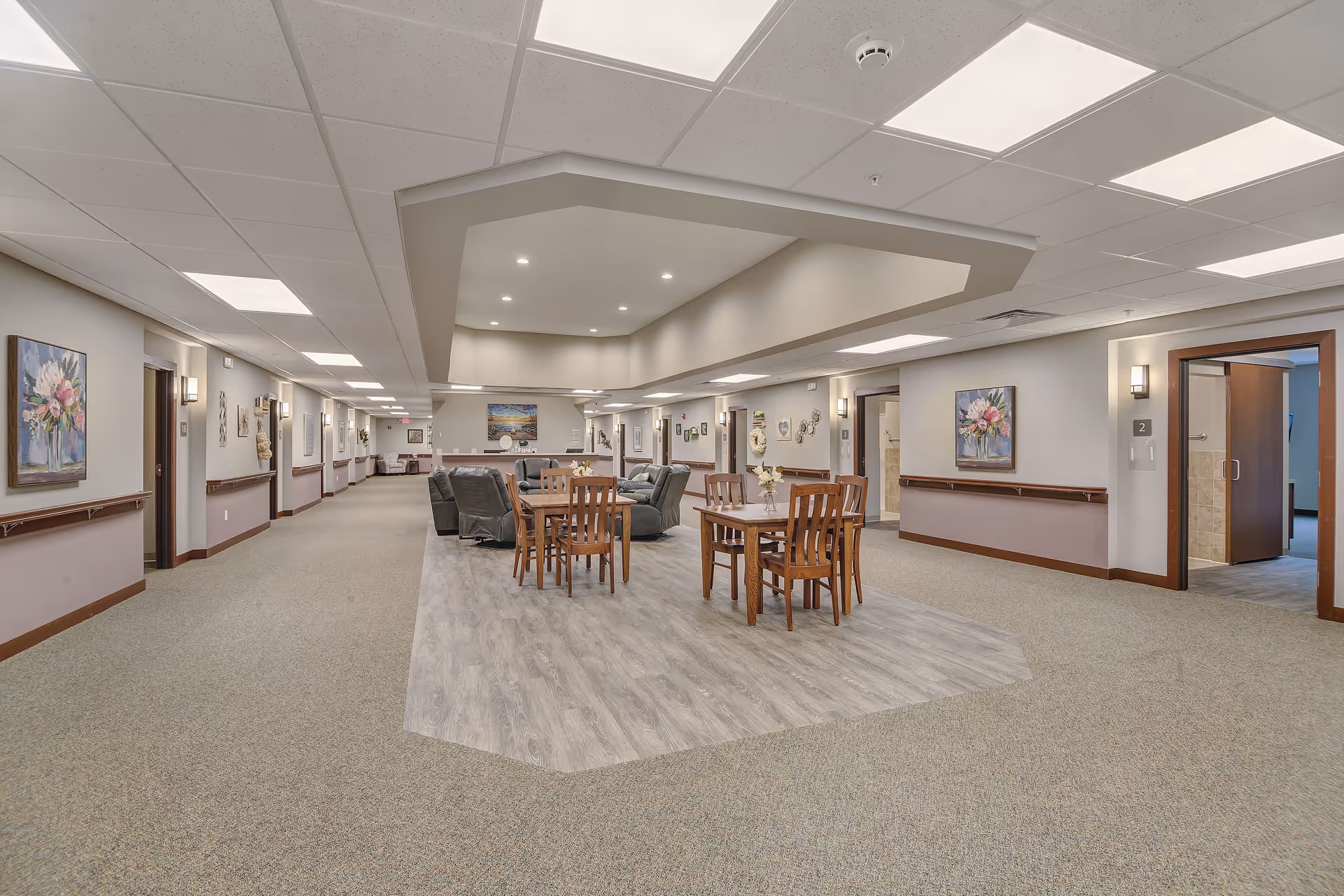 Interior view of a senior living facility hallway with a seating area in the center. The seating area has wooden tables and chairs on a wood-patterned floor section, surrounded by carpeted flooring. Comfortable recliner chairs are arranged near the tables. The walls are decorated with paintings and wall sconces, and several doorways lead to other rooms.