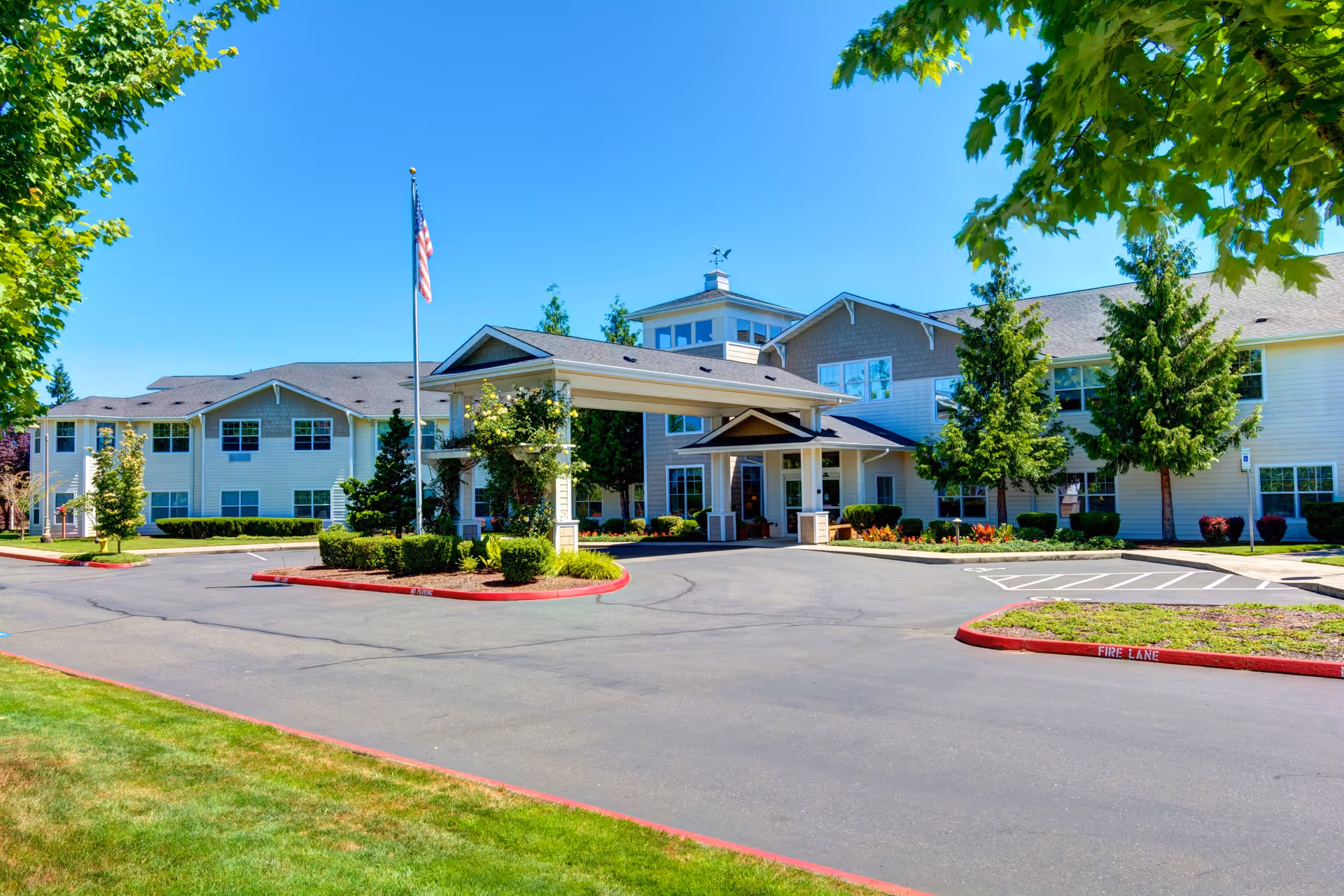Exterior view of Clearwater Springs by Cogir, a two-story senior living facility with a covered entrance, surrounded by trees and landscaping under a clear blue sky.