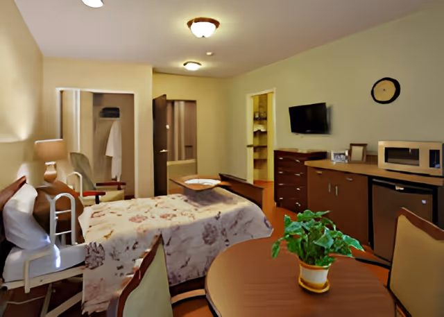 A cozy senior living room with a bed covered by a floral blanket, a bedside table with a lamp, a walker, a round table with a potted plant, chairs, a wall-mounted TV, a microwave, a mini fridge, and a clock on the wall. The room has warm lighting and an open closet with a white robe hanging inside.