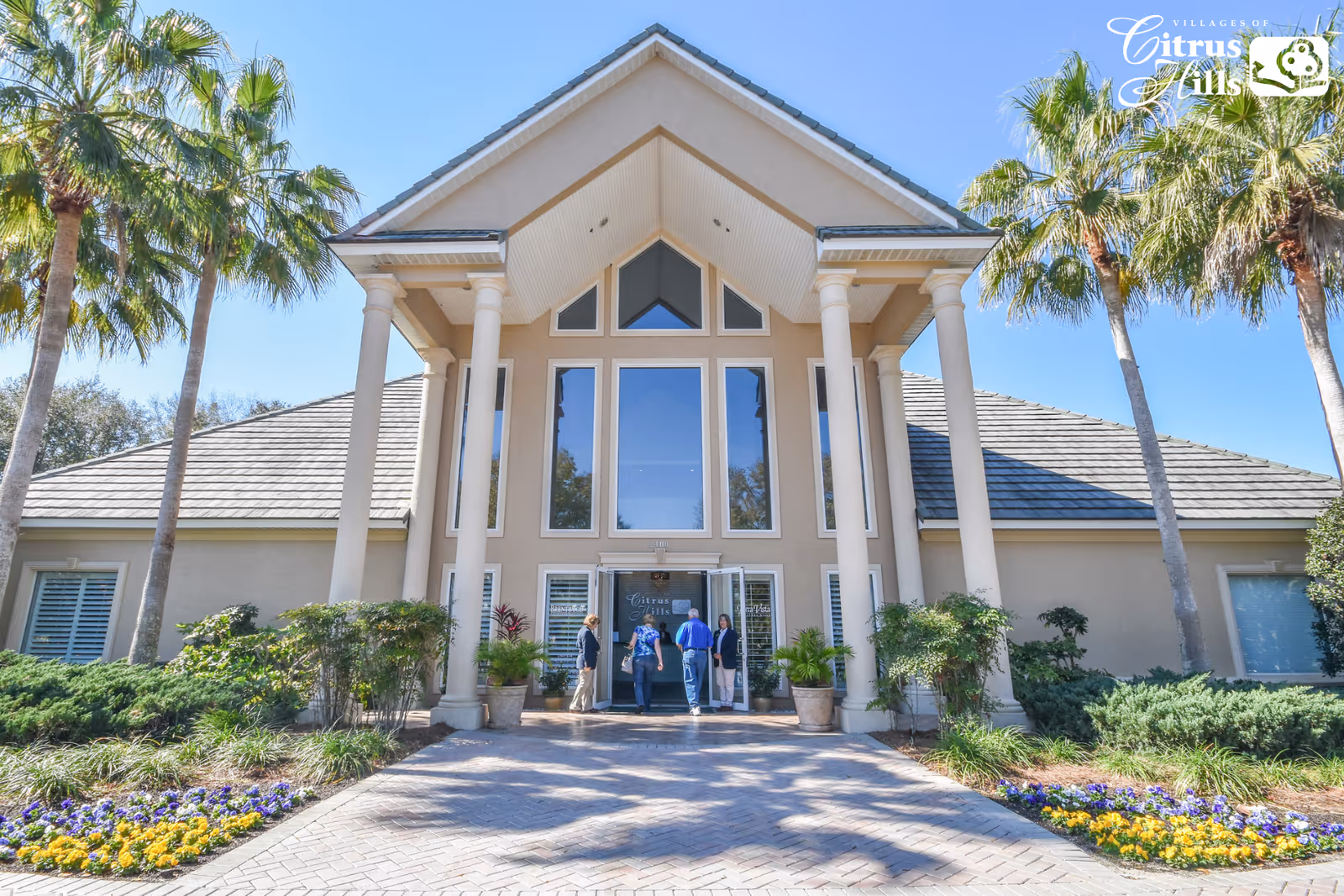 Front exterior view of a large building with tall white columns and large windows. There are palm trees and landscaped flower beds with yellow and purple flowers on either side of the paved walkway leading to the entrance. Four people are standing near the entrance doors. The sky is clear and blue.