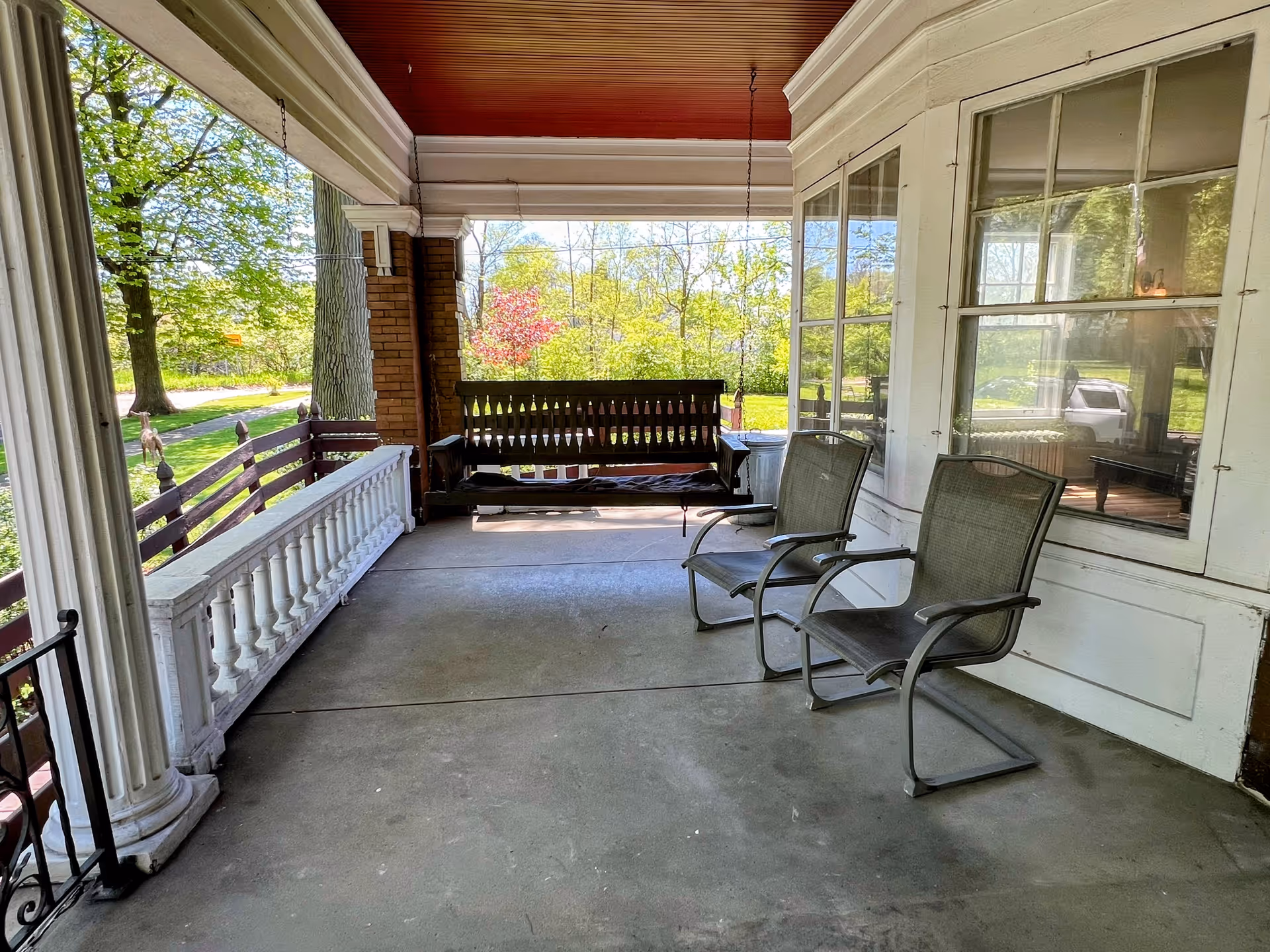 A covered front porch with two metal chairs and a wooden swing overlooking a leafy yard.