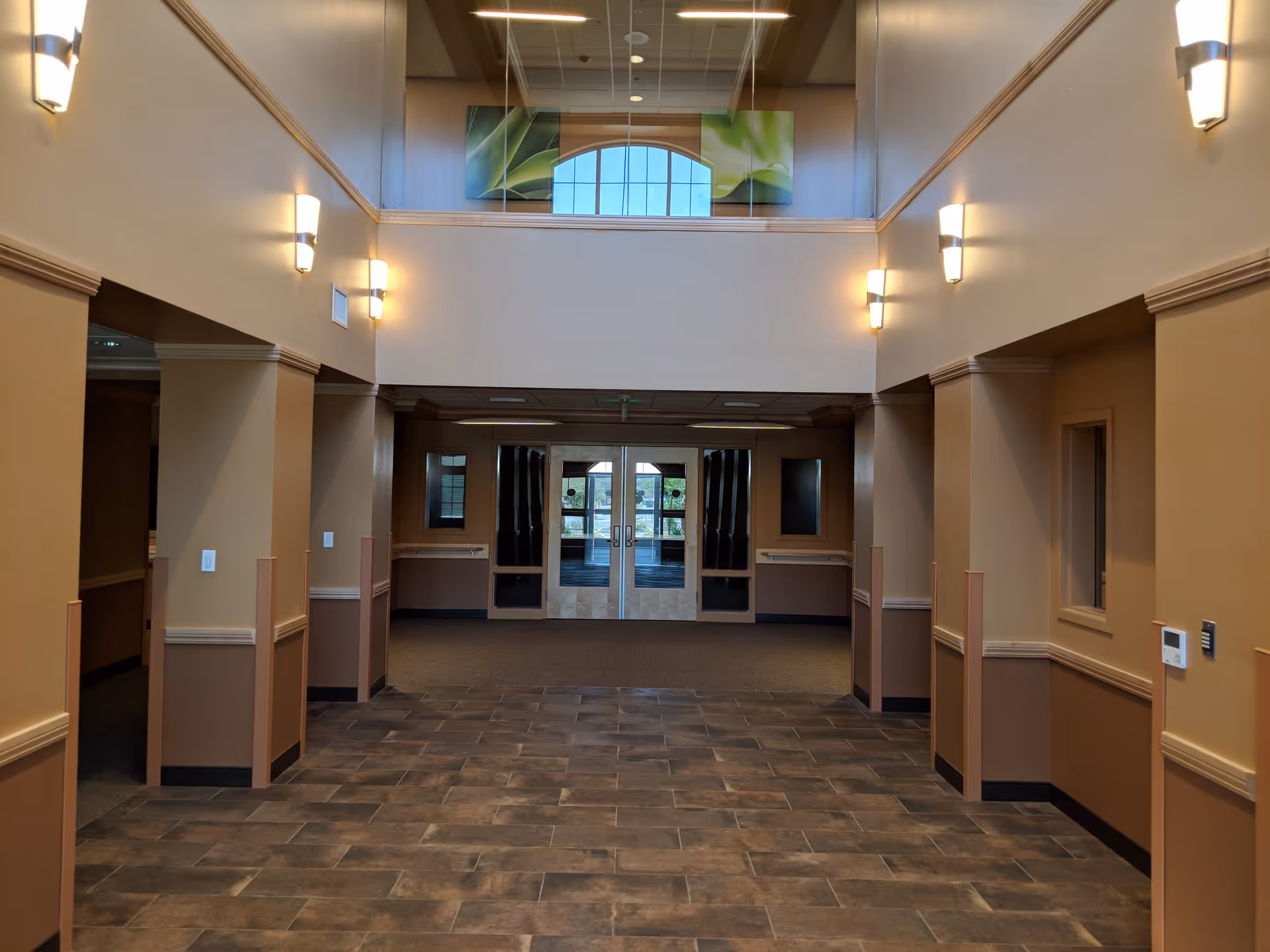 Interior view of a senior living facility hallway with tiled floor, beige and brown walls, wall-mounted lights, and double glass doors at the end. There is a second-floor balcony with glass railing and a large window with green artwork above the doors.