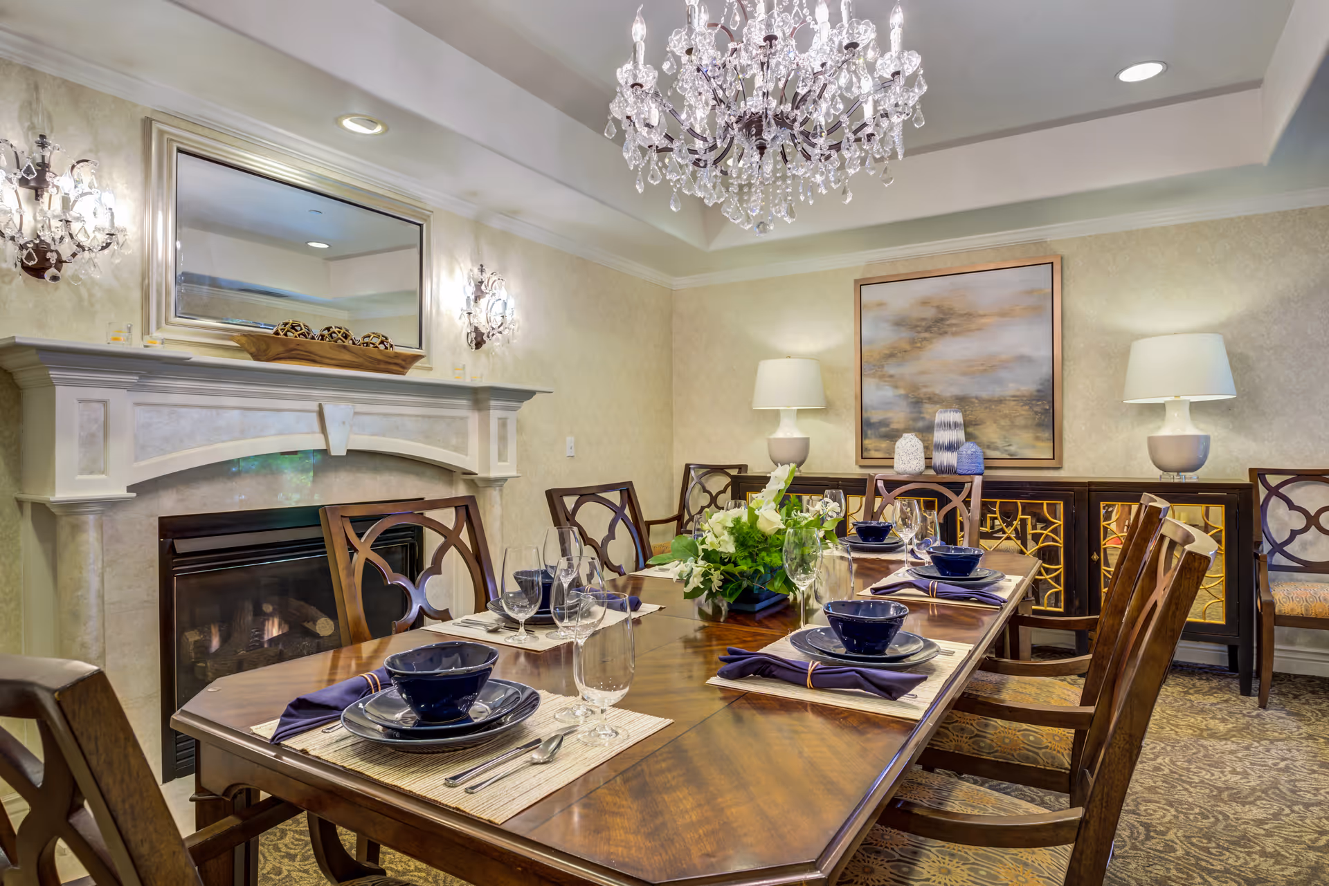 Elegant dining room with a long wooden table set for a meal, crystal chandelier, fireplace, and decorative sideboard with lamps.