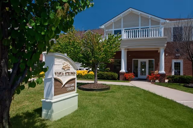 Exterior view of a brick building with white trim and a balcony above the entrance. A well-maintained lawn and landscaping with trees and flowers surround a sign that reads 'Vista Springs Welcome Center'.