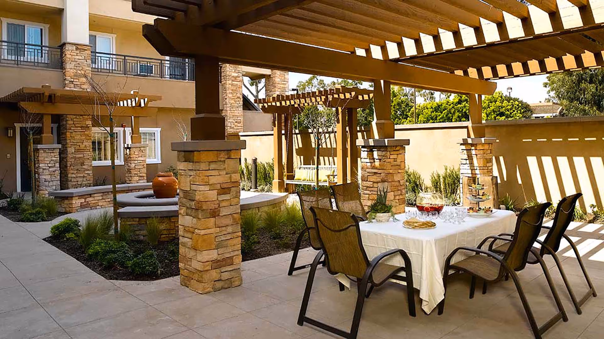 Outdoor patio area with a pergola featuring stone pillars, a table covered with a white tablecloth set with a pitcher of drink, glasses, and snacks, surrounded by six chairs. In the background, there is a swing bench and landscaping with plants and trees.