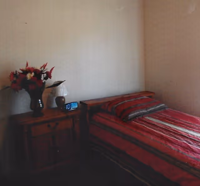 A small bedroom with a red striped bed, wooden nightstand, vase of flowers, lamp, and an alarm clock.