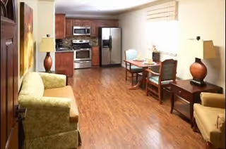 Interior view of a living area with wooden flooring, a green sofa on the left, a small dining table with two chairs near the window, a side table with a lamp, and a kitchen area with stainless steel appliances including a refrigerator, oven, and microwave in the background.