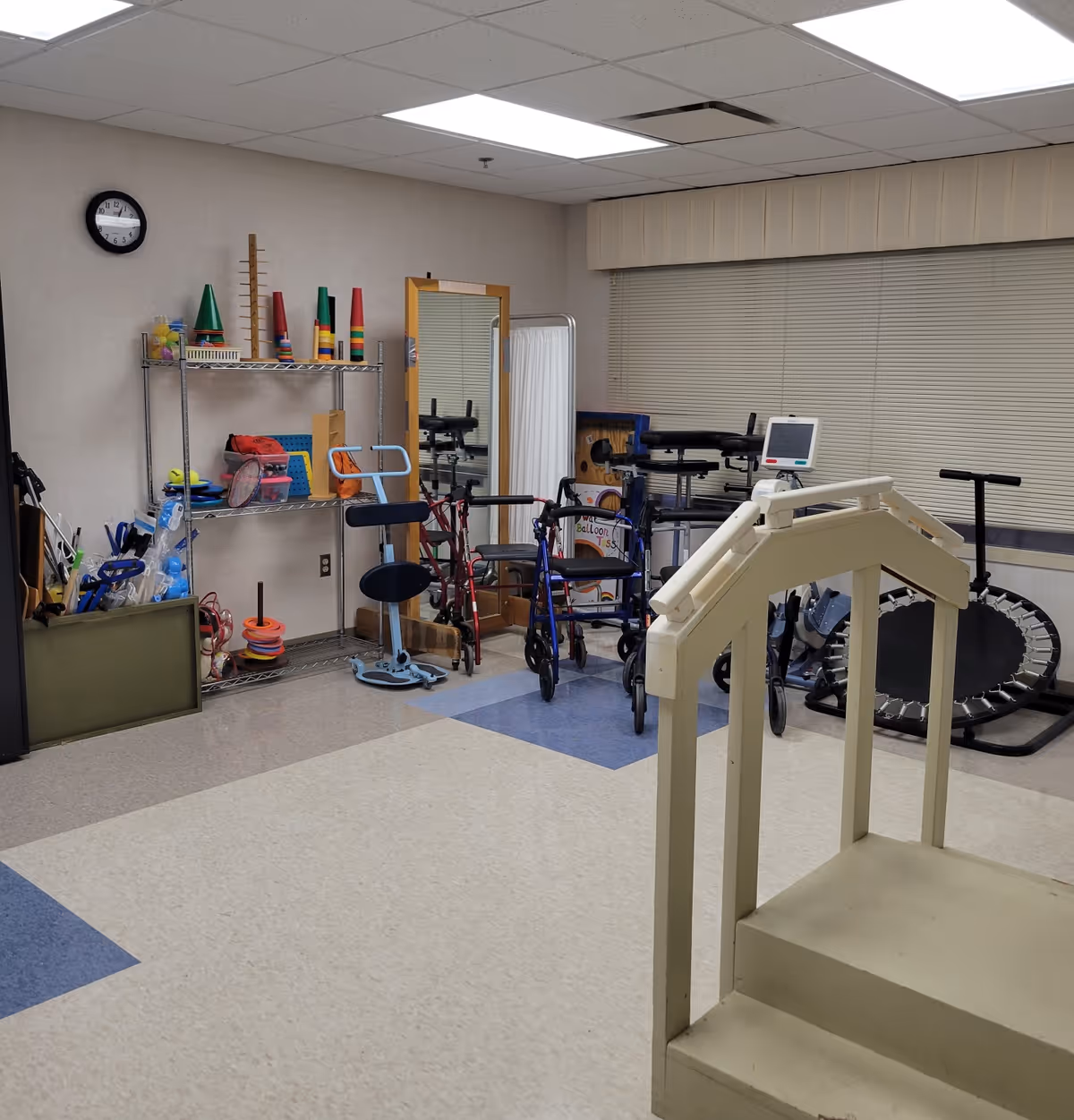 A physical therapy or rehabilitation room with various exercise and mobility equipment including walkers, a small set of stairs with handrails, a mini trampoline, and shelves holding cones, balls, and other therapy tools. There is a large mirror and a clock on the wall.