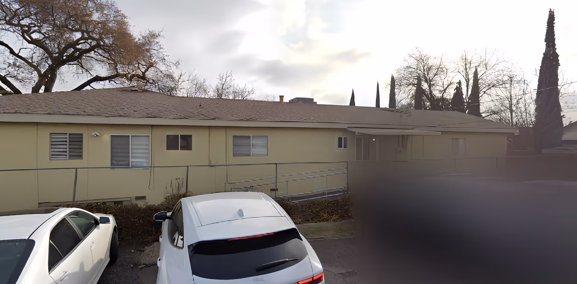 Front exterior of a single-story beige senior living building with a wheelchair ramp and parked cars under an overcast sky.