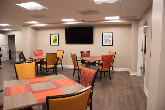 A senior living community common area with multiple square tables and chairs arranged for dining or socializing. The chairs have orange and red patterned upholstery. The room has wood flooring, beige walls, two framed artworks, and a large flat-screen TV mounted on the wall.