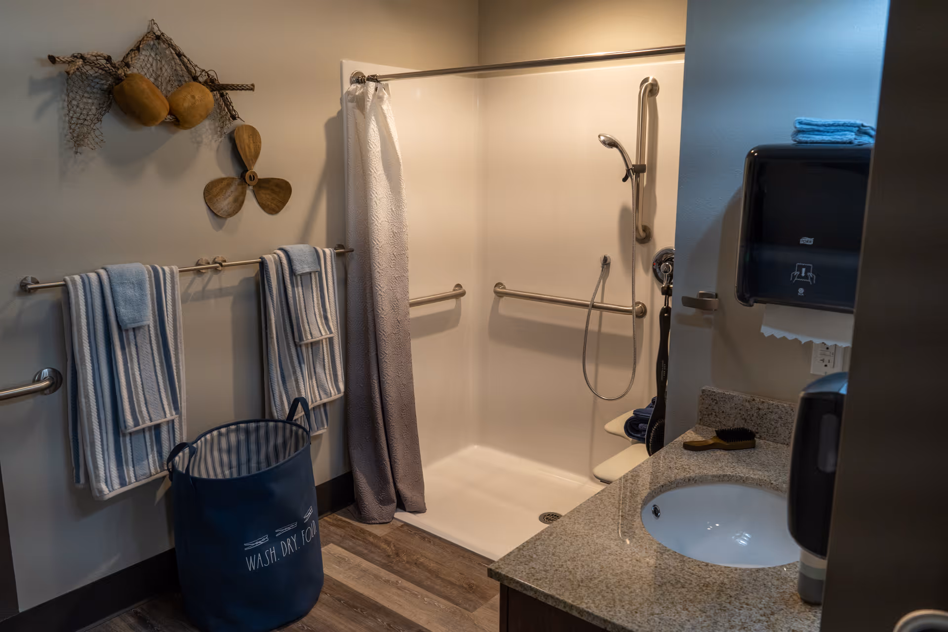 A bathroom with a walk-in shower featuring grab bars and a handheld showerhead. There are two striped towels hanging on a towel rack, a blue laundry basket labeled 'WASH. DRY. FOLD.', a granite countertop with a sink, a soap dispenser, a paper towel dispenser, and decorative wooden items hanging on the wall.
