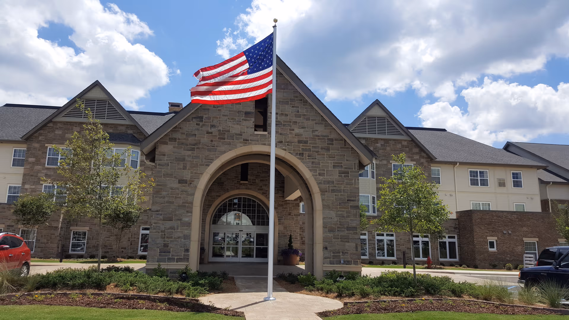 Front exterior view of Cahaba Ridge Retirement Community building with a stone arch entrance, an American flag on a flagpole in front, several small trees, and a partly cloudy sky.
