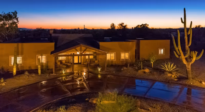 Front entrance of a single-story desert building at dusk with a covered entryway, lit windows, saguaro cacti, and a reflective wet driveway.