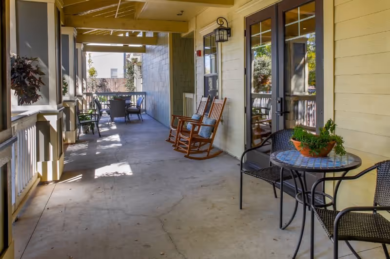 Covered outdoor patio area with rocking chairs, a small round table with two chairs, and a larger dining table with chairs at the far end. The patio has a concrete floor, yellow siding walls, and glass doors leading inside. Potted plants are placed on the tables and railing.
