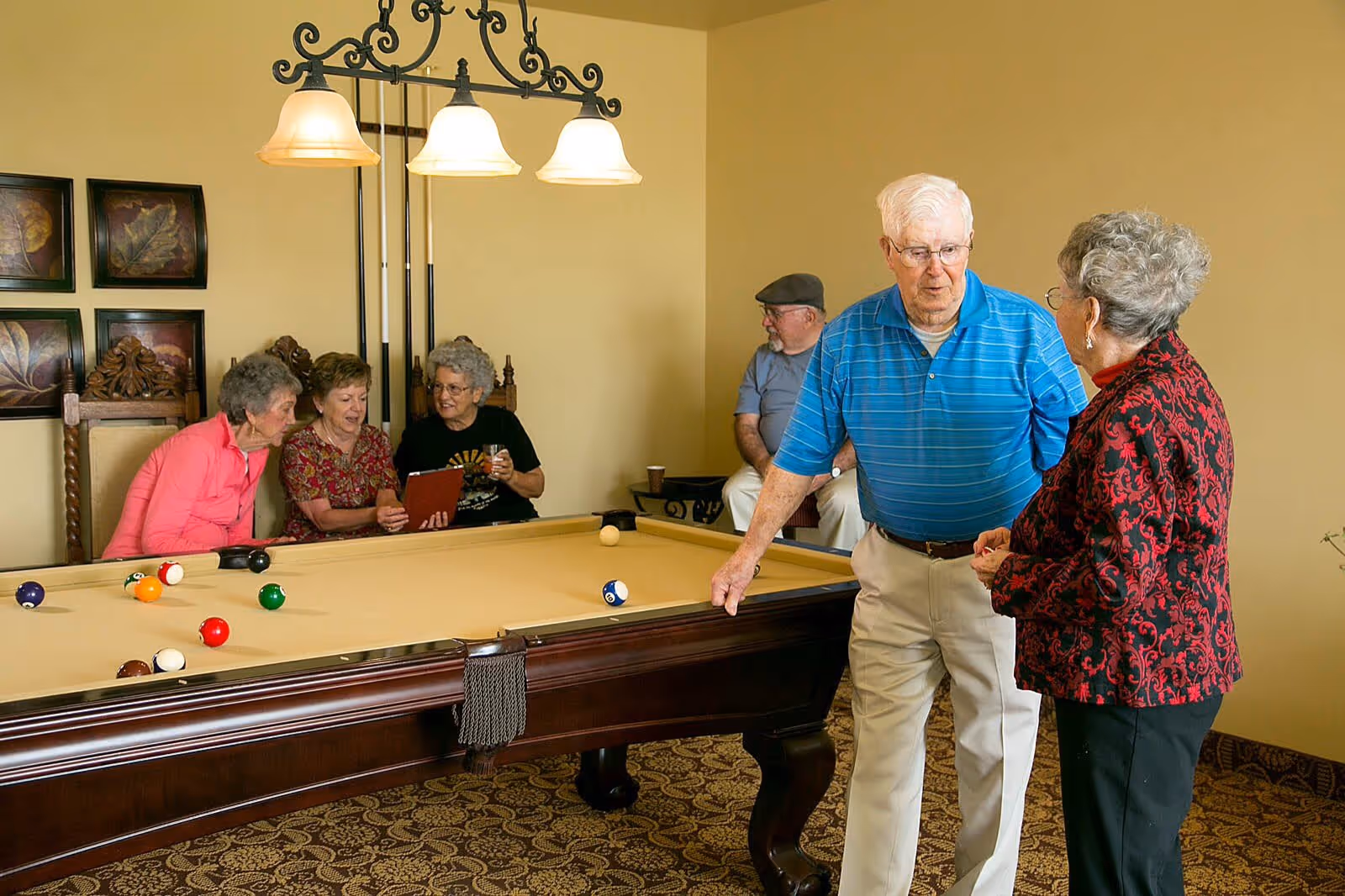 A group of elderly people socializing around a billiards table in a cozy room. Three women are seated on chairs along the wall, one holding a tablet and another holding a drink. An elderly man in a blue striped shirt is standing and talking to a woman in a red and black patterned jacket. Another man wearing a cap is seated in the background. The room has warm yellow walls, decorative framed artwork, and a hanging light fixture above the pool table.