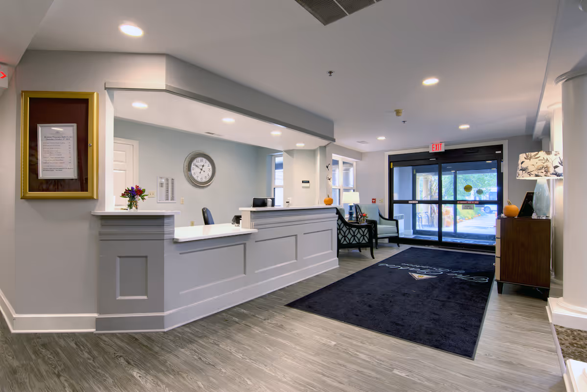 Reception desk and lobby area of a senior living community with seating, an entrance door, and a dark floor mat.