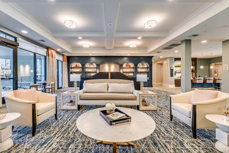 A spacious and well-lit living room area with a beige sofa and two matching armchairs arranged around a round marble coffee table. The room features a patterned carpet, built-in dark wood bookshelves with decorative items, and large windows with curtains allowing natural light to enter. There are modern ceiling lights and side tables with lamps beside the sofa.
