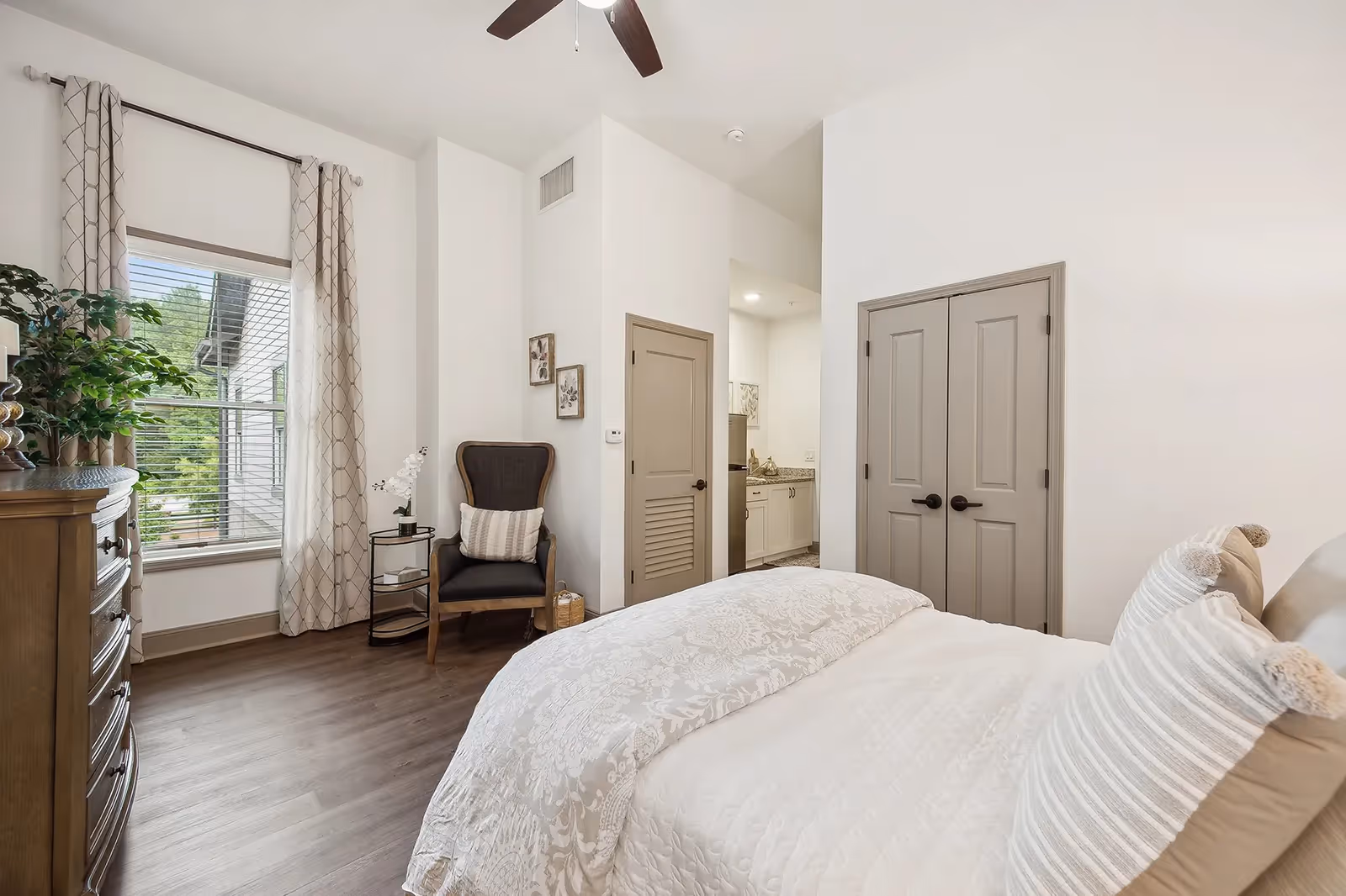 Bright furnished bedroom with a bed, armchair, dresser, window with curtains, and double closet doors.