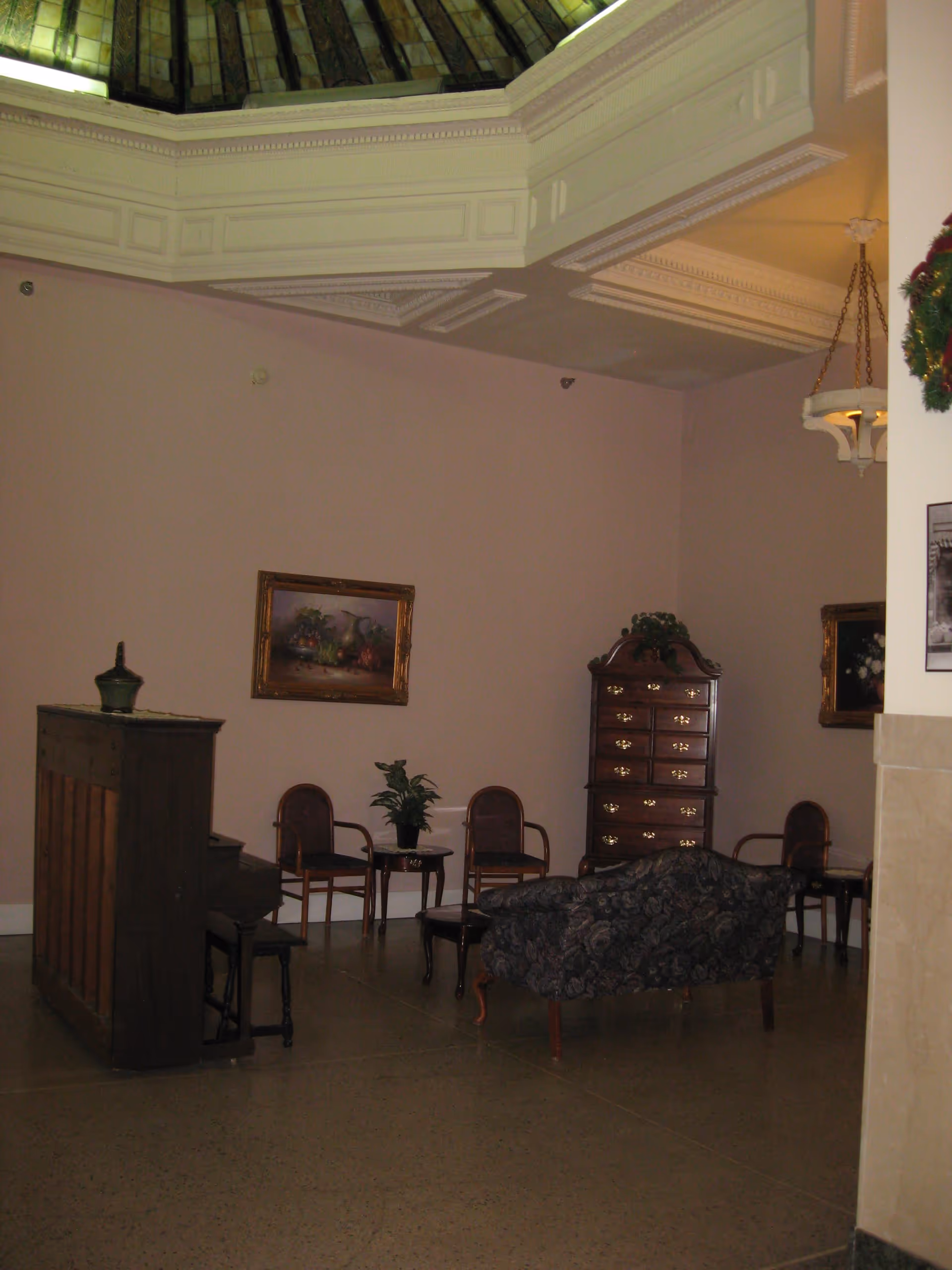 Interior room with a high ceiling and decorative molding, featuring a wooden upright piano, a floral-patterned loveseat, several wooden chairs, a small table with a plant, a tall chest of drawers, and framed paintings on the walls.