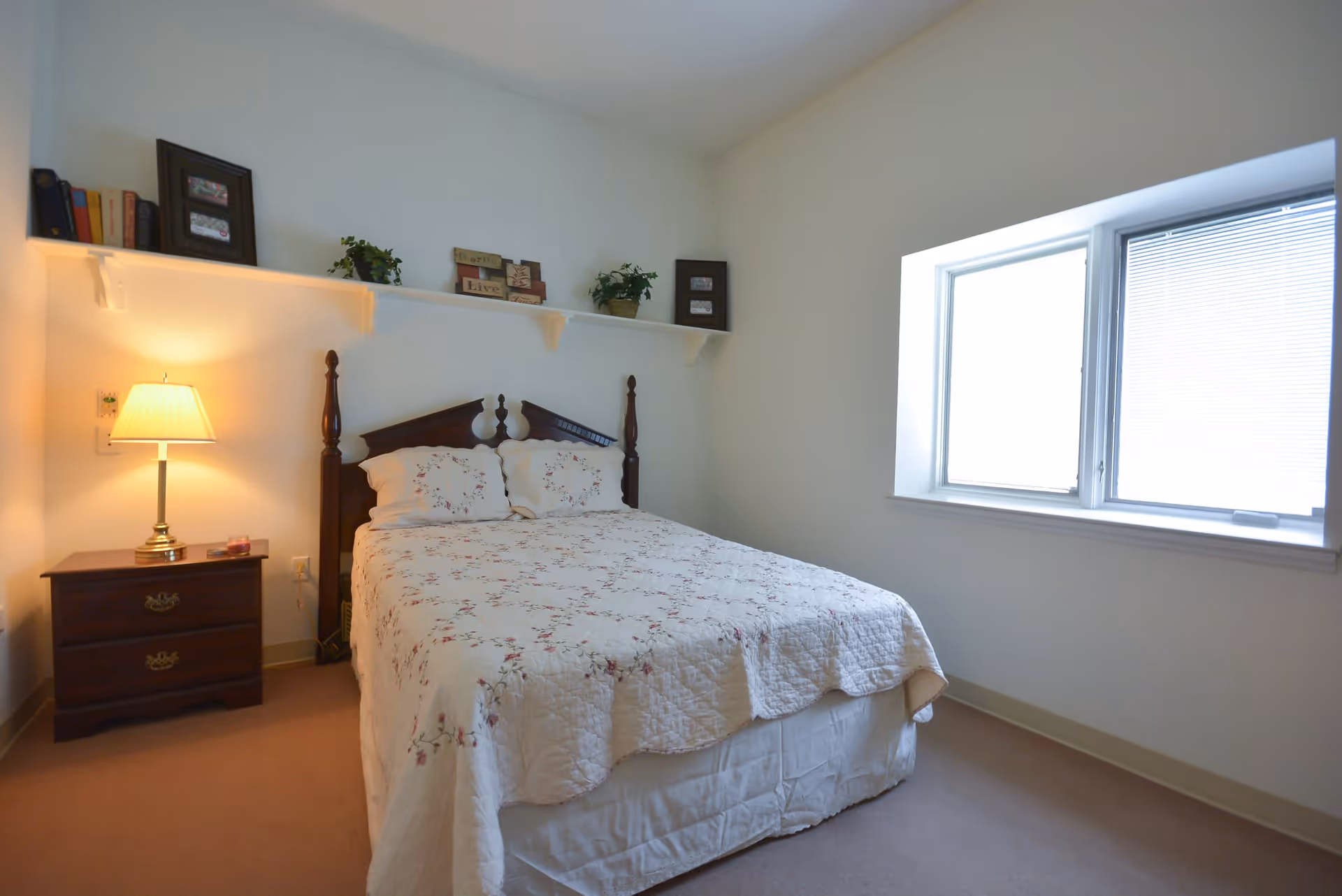 A simple bedroom with a double bed covered in a floral quilt and matching pillows. There is a wooden nightstand with a lamp and a candle on it next to the bed. Above the bed, a white shelf holds books, framed pictures, and small potted plants. A window with blinds is on the right wall, letting in natural light.