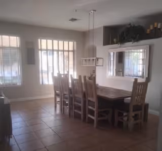 A dining room with a wooden dining table and six matching wooden chairs. The room has tiled flooring, large windows with vertical blinds allowing natural light to enter, a rectangular hanging light fixture above the table, and a large mirror on the wall reflecting part of the room. There are decorative items on a shelf above the mirror.