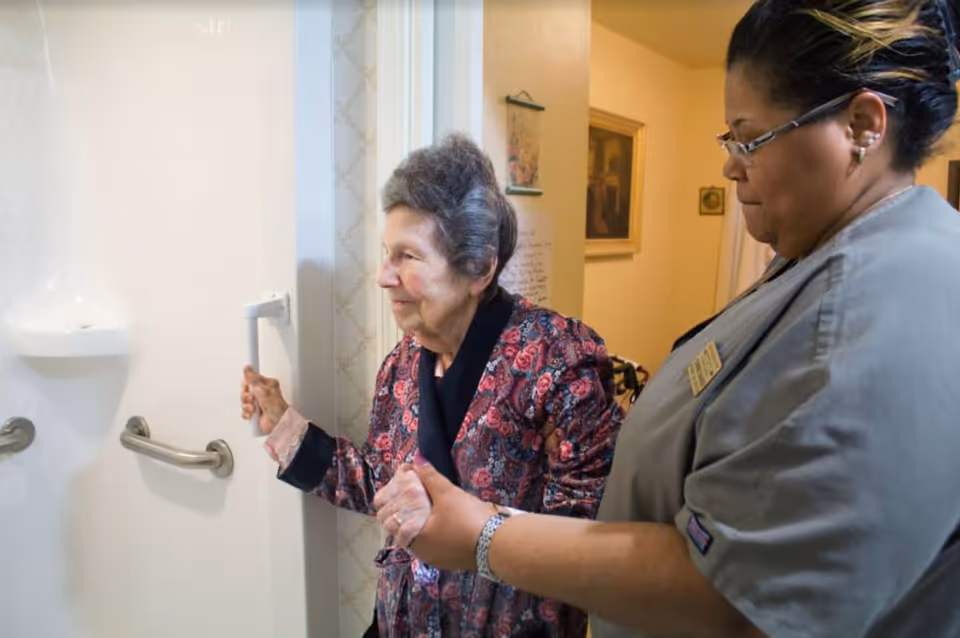 An elderly woman holding onto a grab bar inside a shower while being assisted by a caregiver in a gray uniform inside a senior living facility.