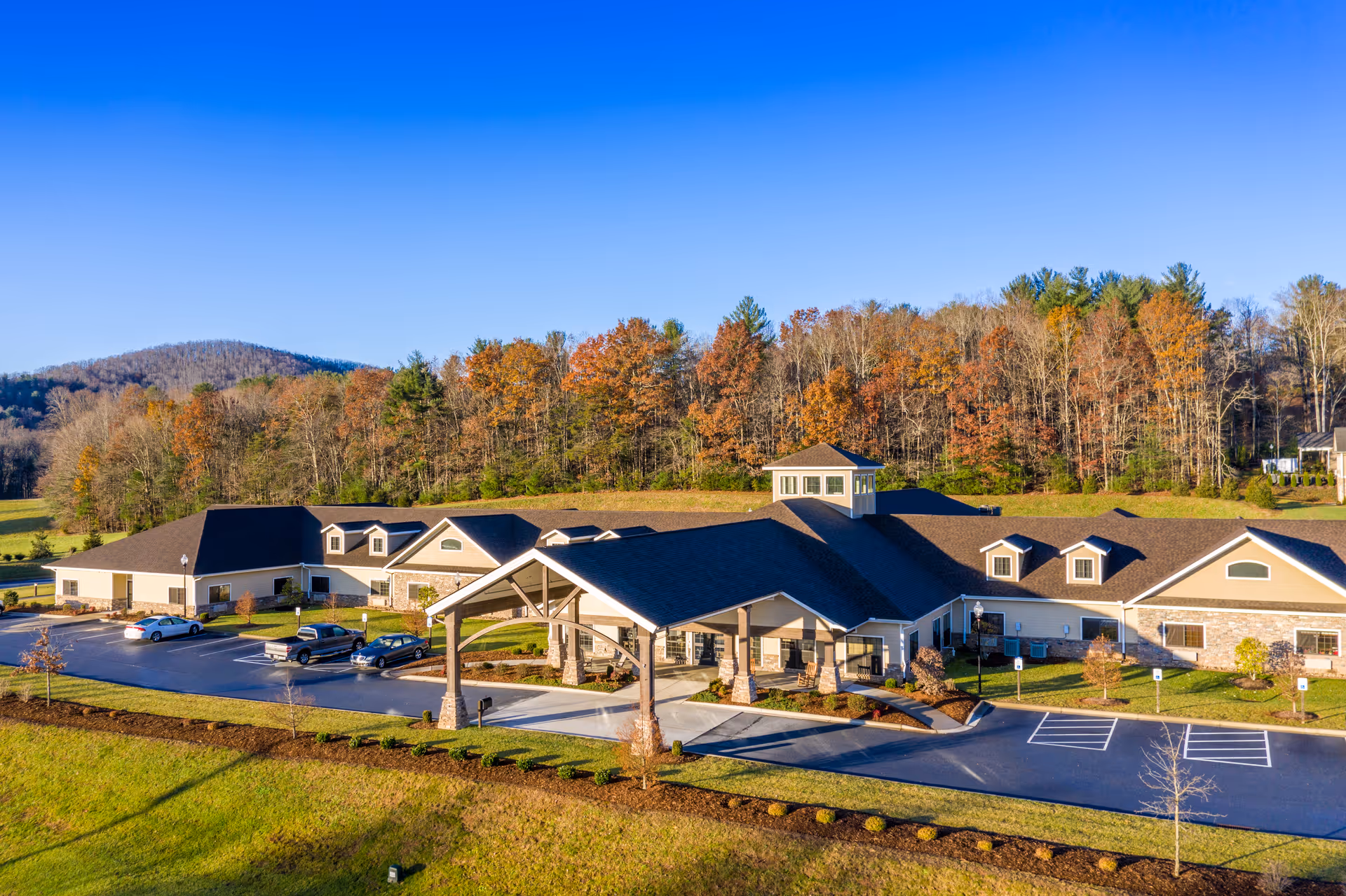 Aerial view of The Lodge at Mills River Health and Rehabilitation building surrounded by a parking lot and landscaped greenery, with a backdrop of trees showing autumn foliage and a clear blue sky.