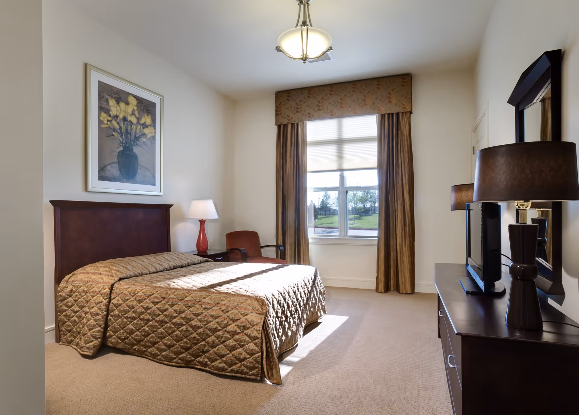 A well-lit bedroom with a large window covered by curtains and a valance. The room features a bed with a quilted bedspread, a wooden headboard, a nightstand with a lamp, a chair, a dresser with a TV and a lamp, and a framed floral painting on the wall.