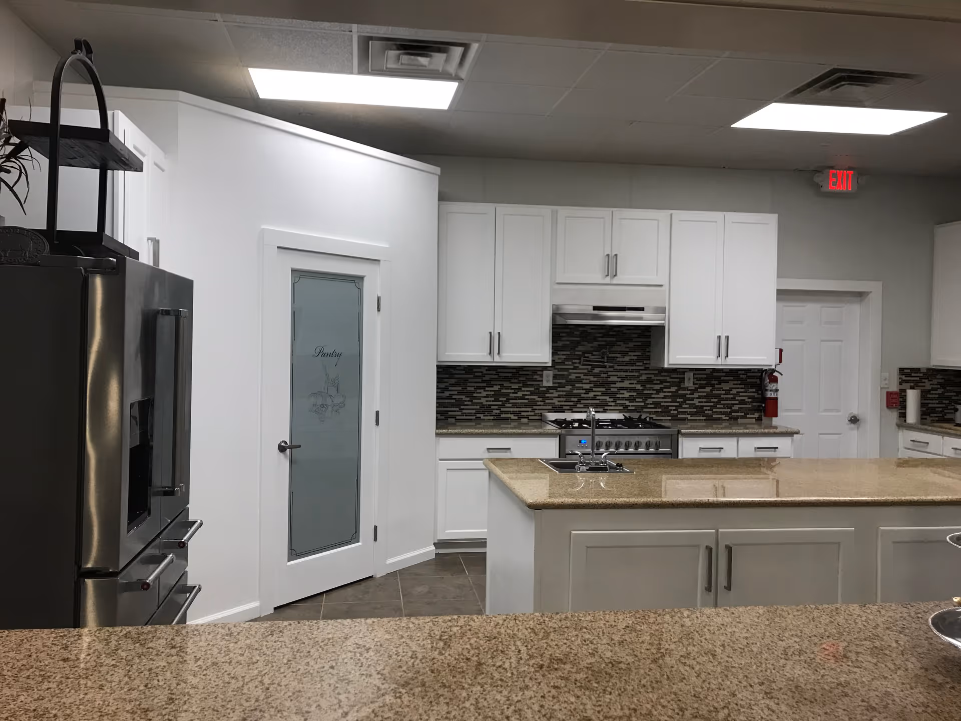 Communal kitchen with white cabinets, a granite island and stainless steel appliances including a refrigerator and stove, with a frosted pantry door.