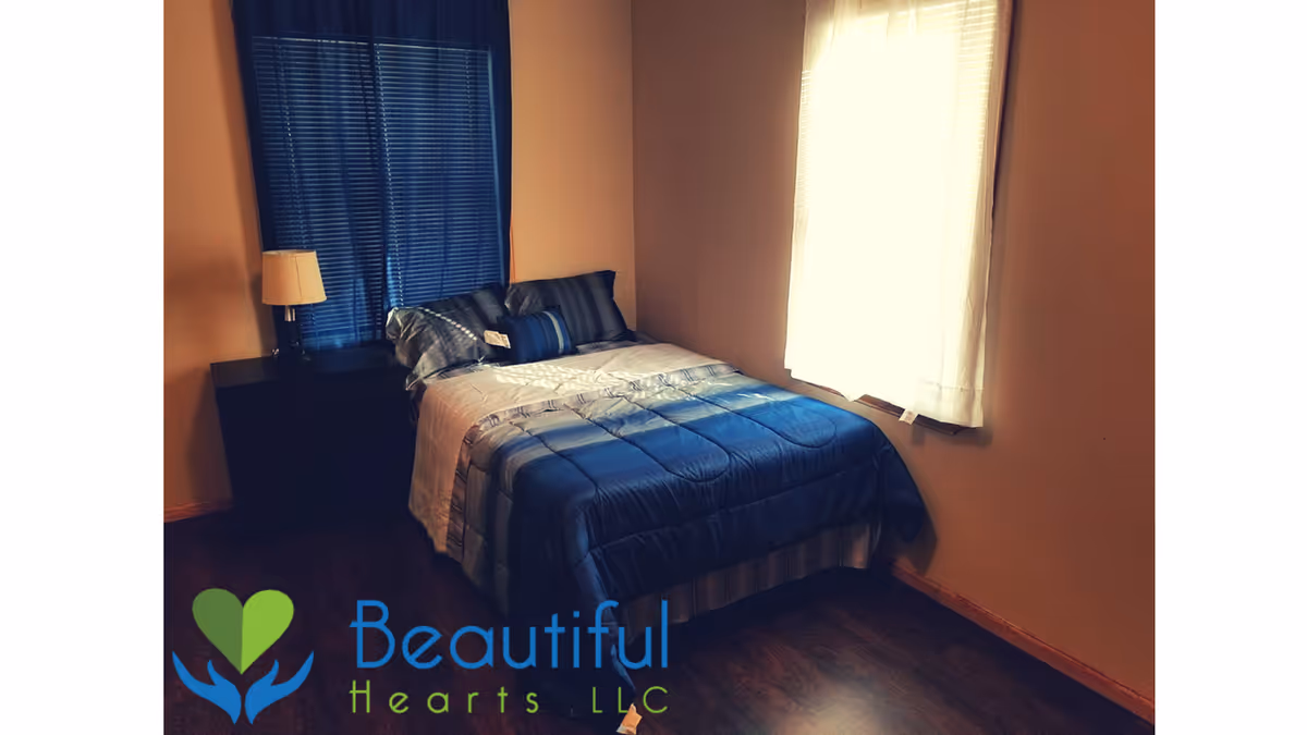 Small bedroom with a twin bed made with blue bedding, a nightstand and lamp, and two windows with curtains.