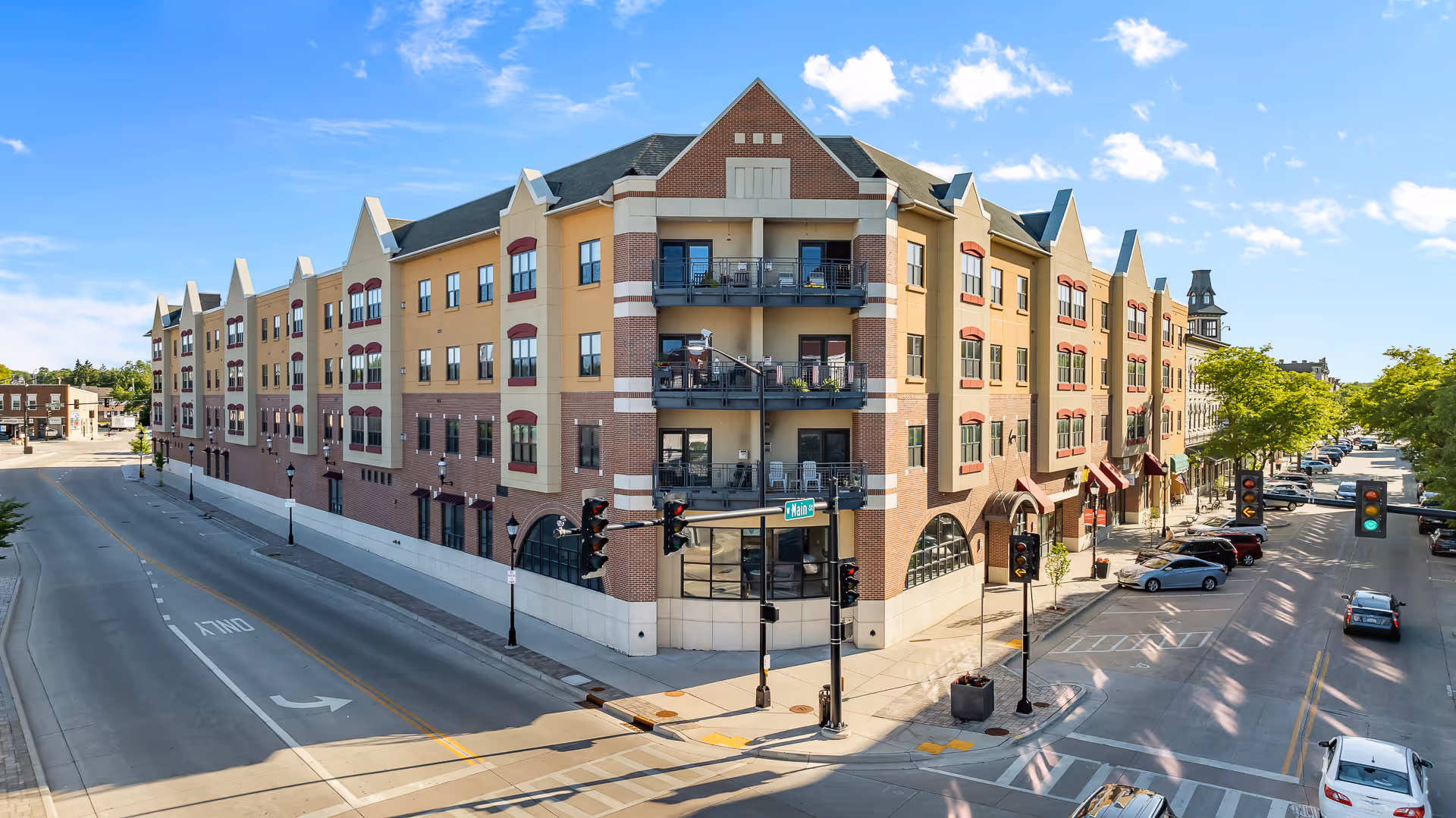 Exterior view of a multi-story senior living facility building on a street corner with balconies, windows, and traffic lights under a blue sky with some clouds.