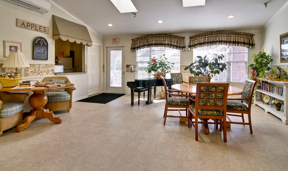 A bright and cozy common area in a senior living facility featuring a round wooden table with four floral upholstered chairs, a black grand piano near large windows with plaid valances, several potted plants, a small seating area with a round wooden table and cushioned chairs, and a white bookshelf filled with books and decorative items.