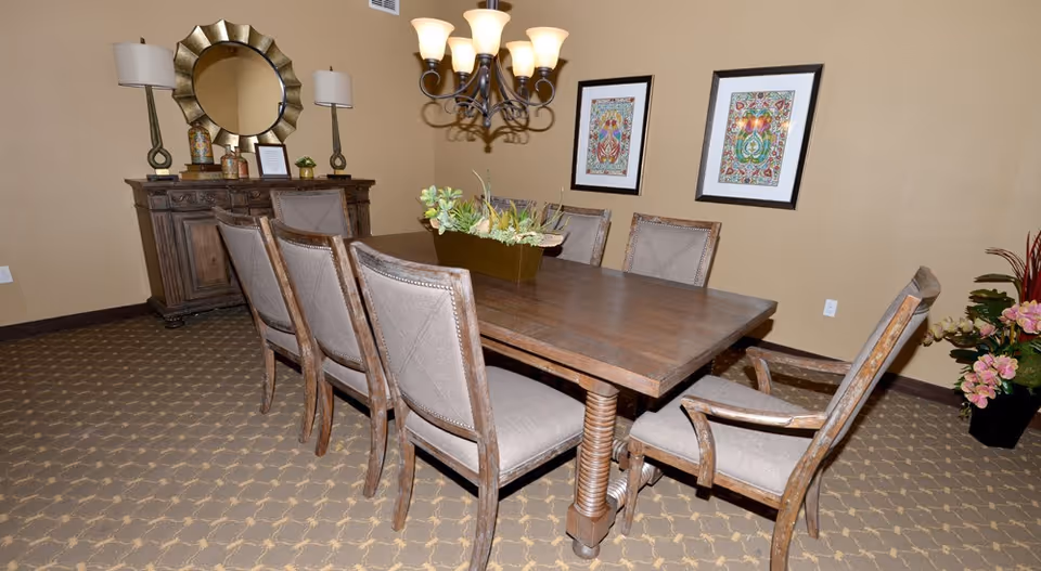 A dining room with a large wooden table surrounded by eight upholstered chairs. The table has a rectangular planter with green plants as a centerpiece. On the wall behind the table are two framed colorful artworks. A wooden sideboard with decorative items and a round mirror is against the far wall. Two table lamps and a chandelier with multiple lights hang above the table. The room has beige walls and patterned carpet flooring.