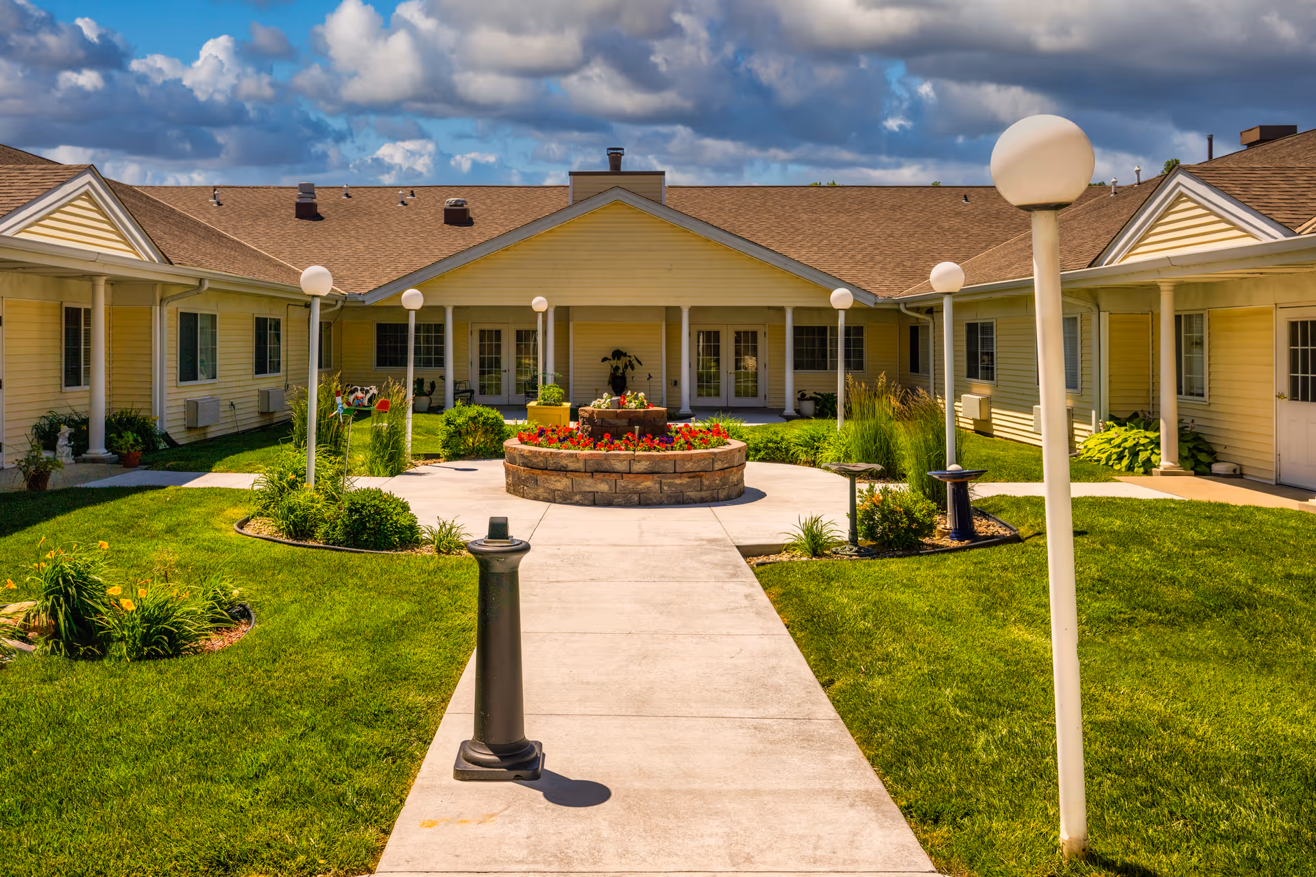 Outdoor courtyard area of a senior living facility with a central circular flower bed surrounded by a concrete walkway, green grass, and lamp posts. The building is a single-story structure with beige siding and a brown roof under a partly cloudy sky.