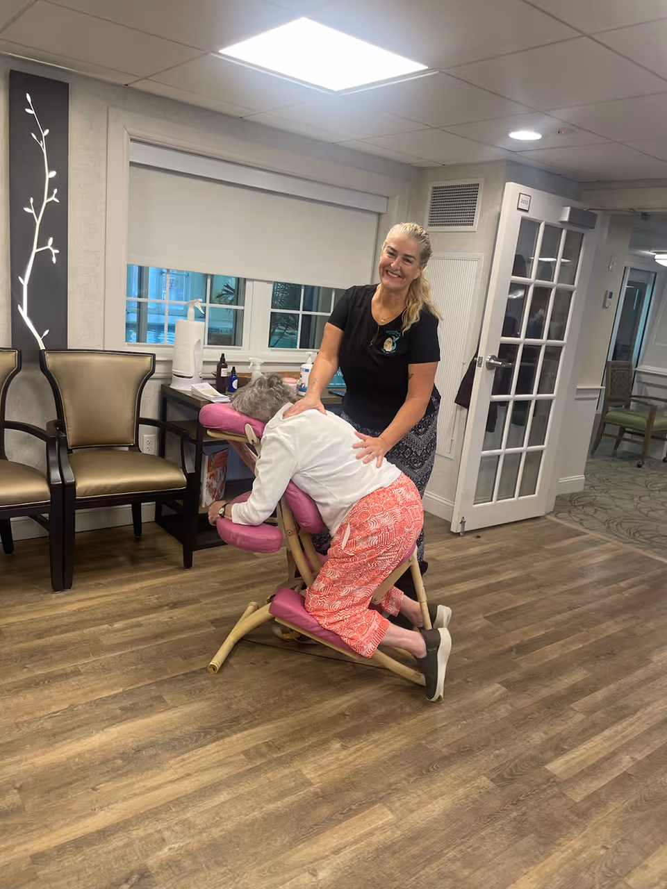 A woman is giving a seated massage to an elderly woman who is kneeling on a specialized massage chair in a well-lit room with wooden flooring. There are two chairs against the wall, a window with a white blind, and a door with glass panels in the background.