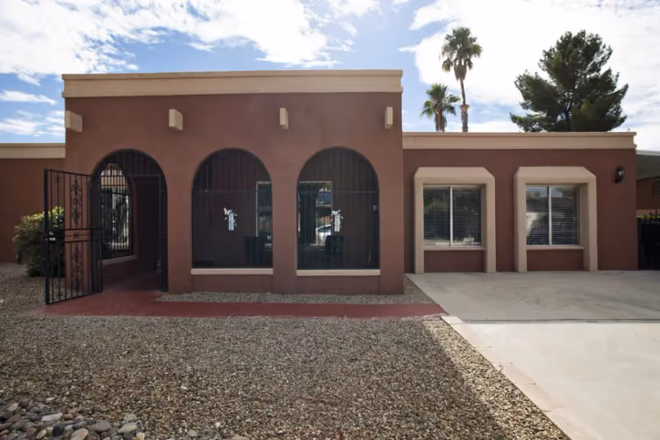 Exterior view of a single-story building with a brown facade featuring three arched windows with black metal bars and two rectangular windows with white blinds. There is a gravel-covered front yard and a concrete driveway. Palm trees and other greenery are visible in the background under a partly cloudy sky.