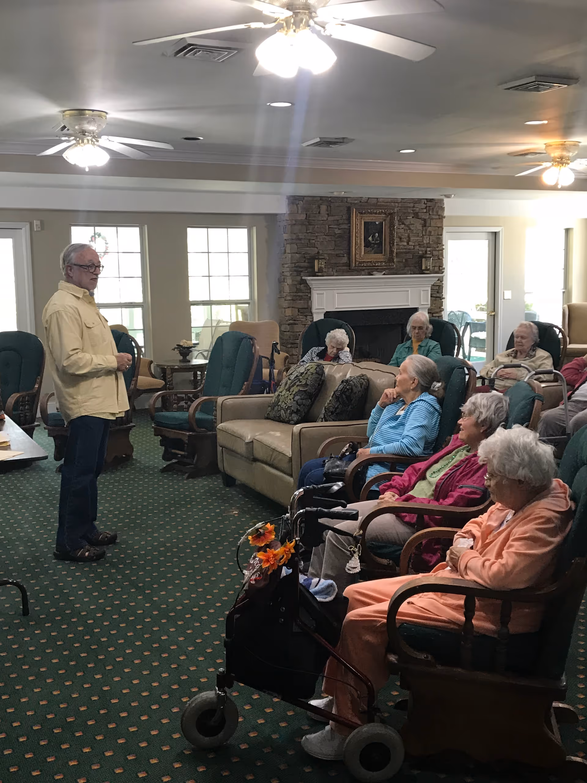 A group of elderly people seated in a common living area with green carpet and ceiling fans. A man stands in front of them, possibly speaking or leading an activity. The room has large windows, a stone fireplace with a framed picture above it, and comfortable chairs and sofas arranged around the space.